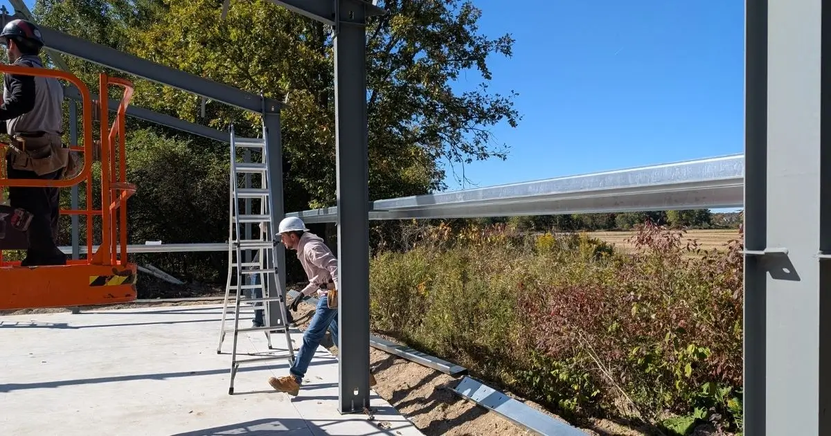 Two construction workers assembling a steel building frame on concrete foundation in Ontario under clear blue sky